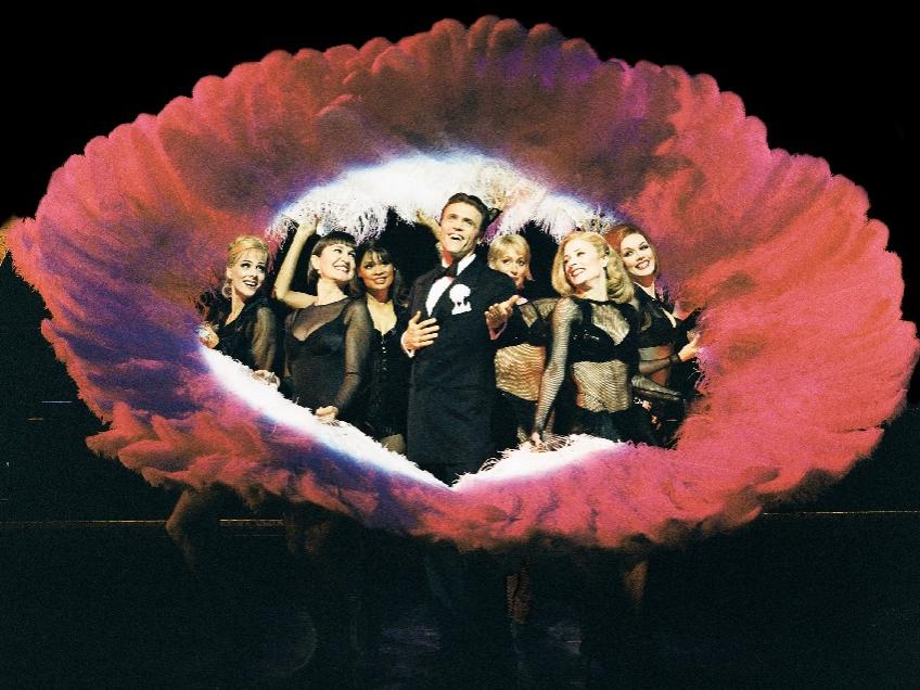 A male actor representing lawyer Billy Flynn stands in the center of a circle of feather fans help by six woman actors, who surround him.