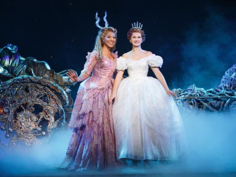 A fairy godmother wears a crown and full-skirted dress while she gestures and smiles at Cinderella in her sparkling dress.