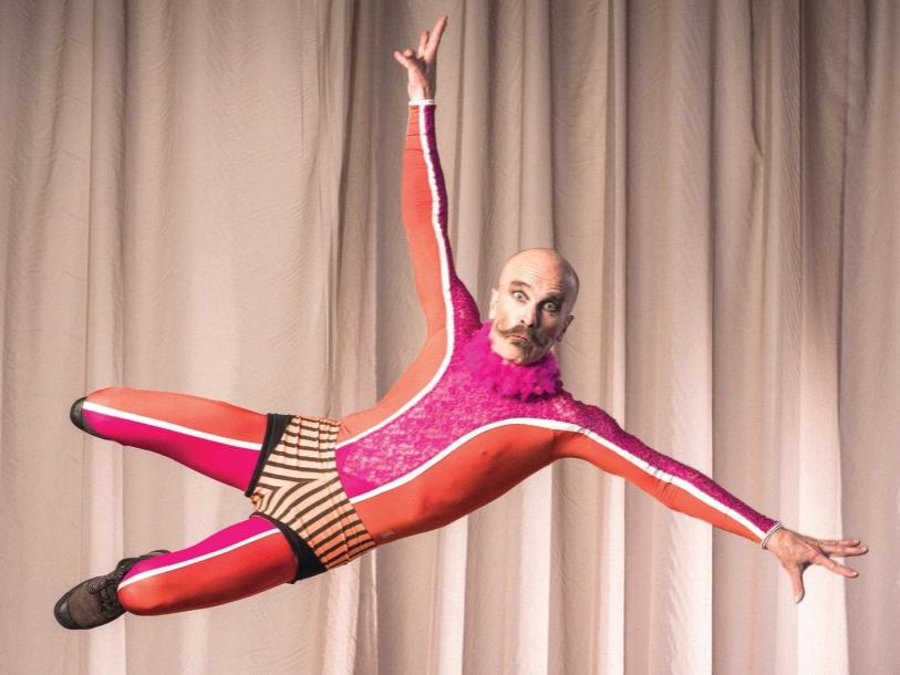 A Circus Oz acrobat is seen suspended in air with his arms extended and his legs leaning toward to his right.