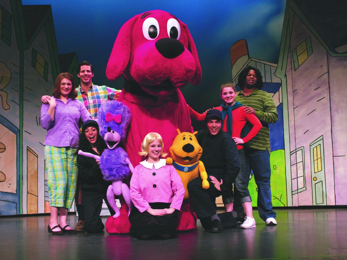 Clifford the Big Red Dog and his three companions dance on stage in front of a drawing of a neighborhood.