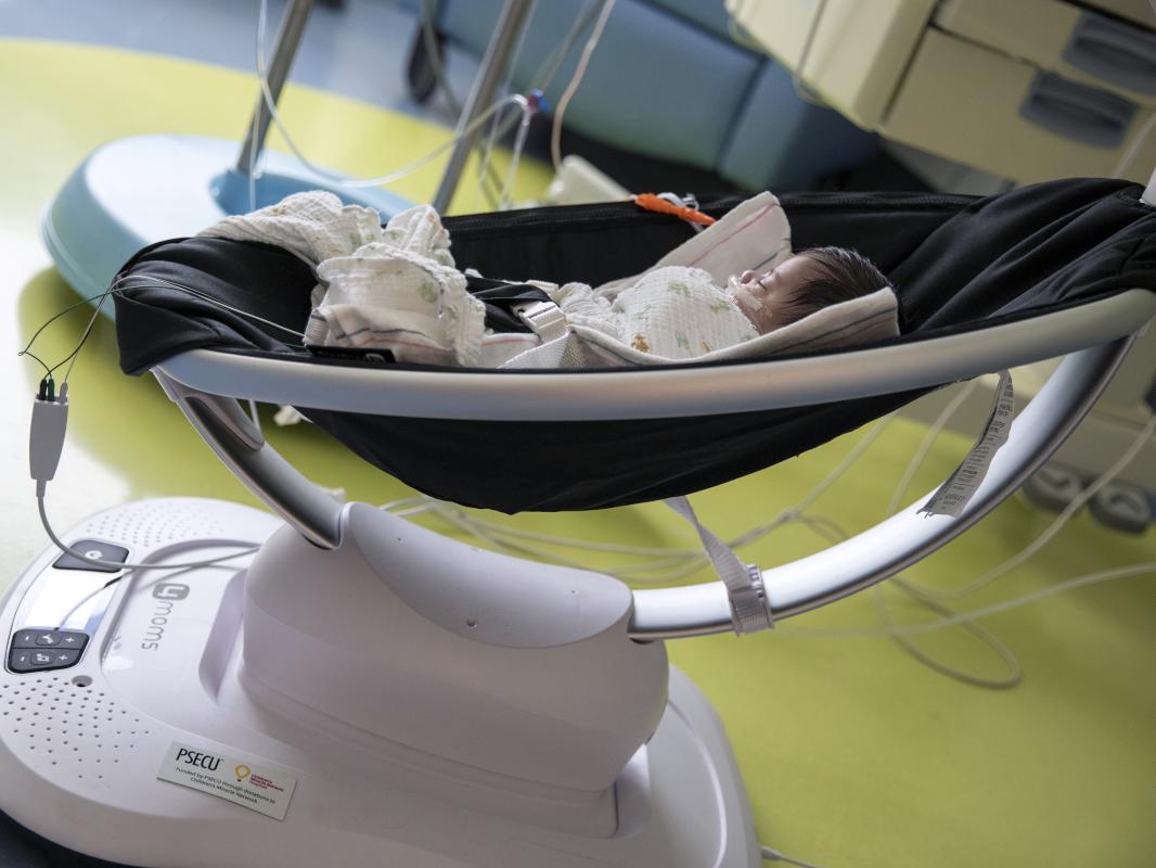 An infant takes a nap in a Mamaroo device, which is resting on the floor of a hospital room.