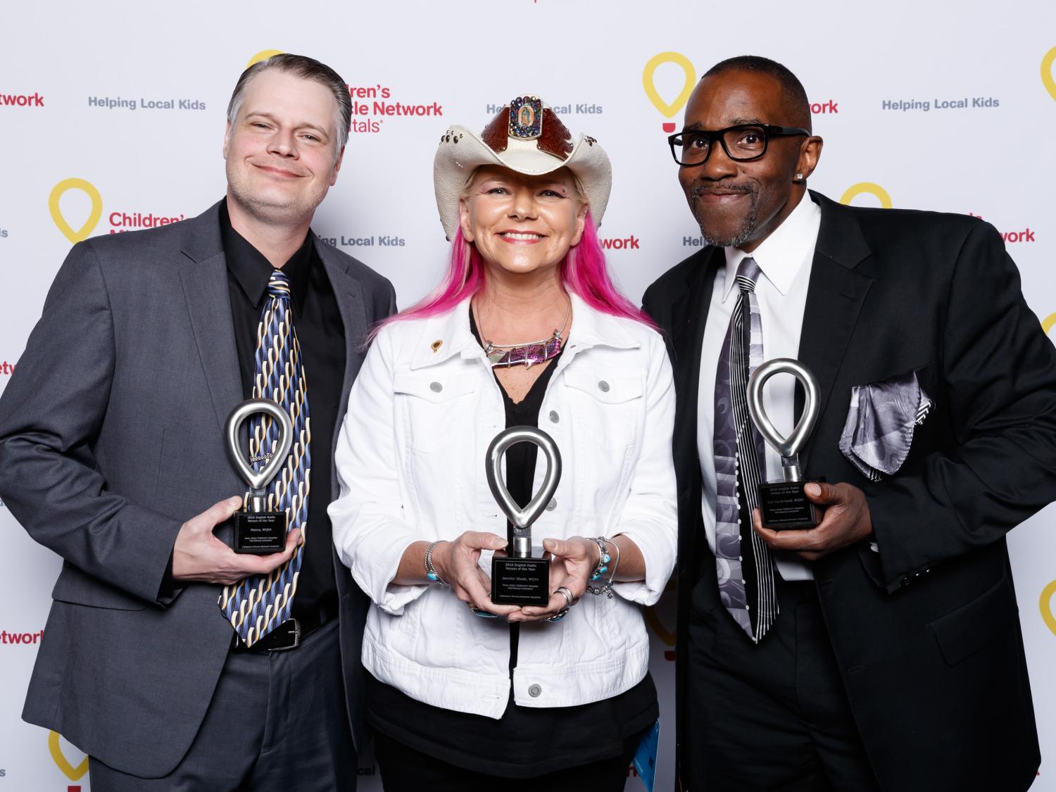Nipsey, Jen Shade and Earl David Reed pose for a photo while holding small trophies, standing in front of a backdrop that has several CMN Hospitals logos.