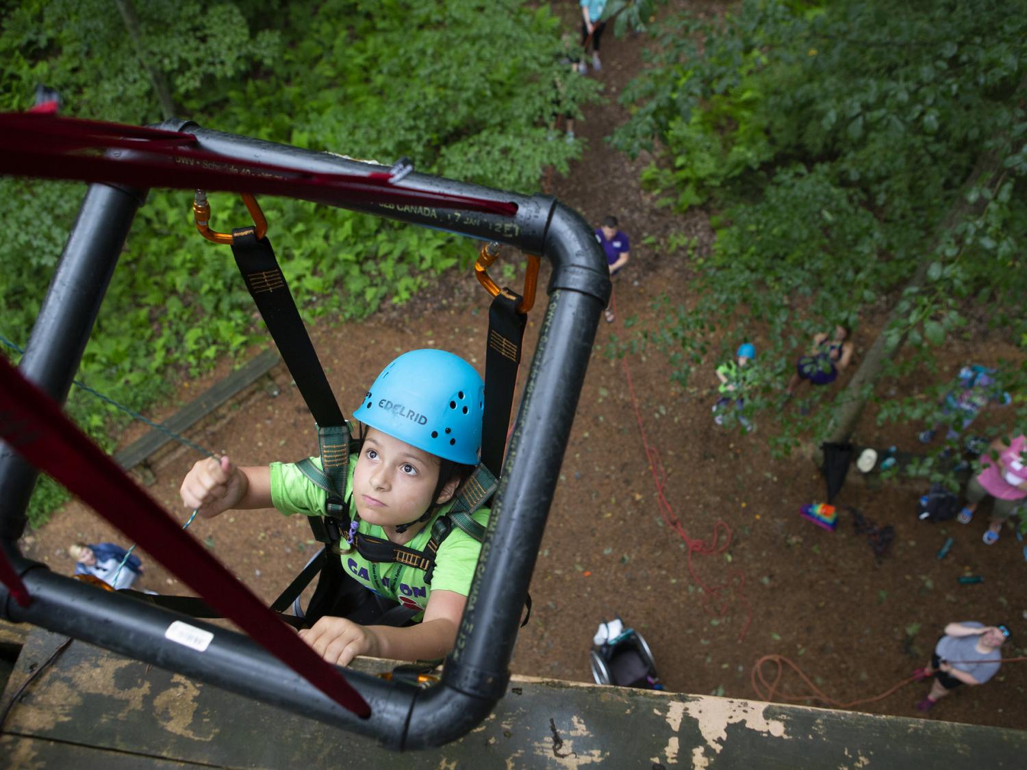 View from the top of a rock wall, looking down, as a young girl in climbing gear and supported by a square metal frame climbs the wall. About 6 people are on the ground, looking up.