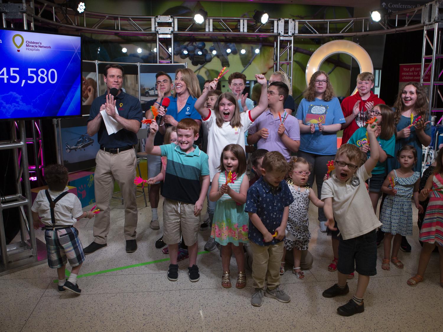 Several people -- more than a dozen children and two adults -- share space on a TV set. The Children's Miracle Network logo is in the background on the right; a TV monitor with the figure "$4,445,580" is on the left.