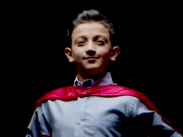 A young boy in a blue shirt and red cape looks into the camera, posing in front of a dark background.