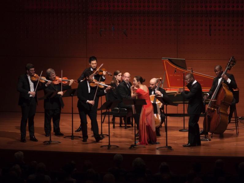 A group of formally dressed musicians perform on classical instruments while standing on a stage.