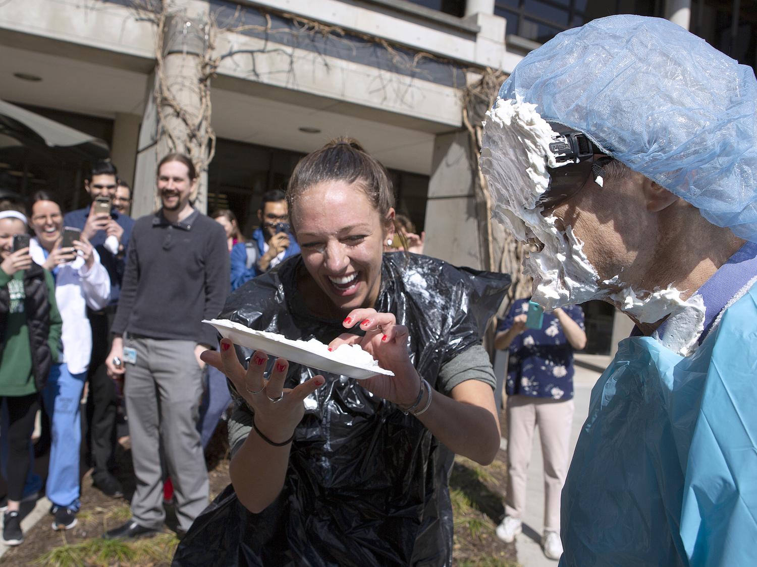 Dr. Michael Katzman, stands with whipped cream on his face and sunglasses. He is wearing a paper smock and hair net. First-year medical student Madelaine Fritsche laughs and holds a plate with whipped cream. Behind them is a large group of onlookers.