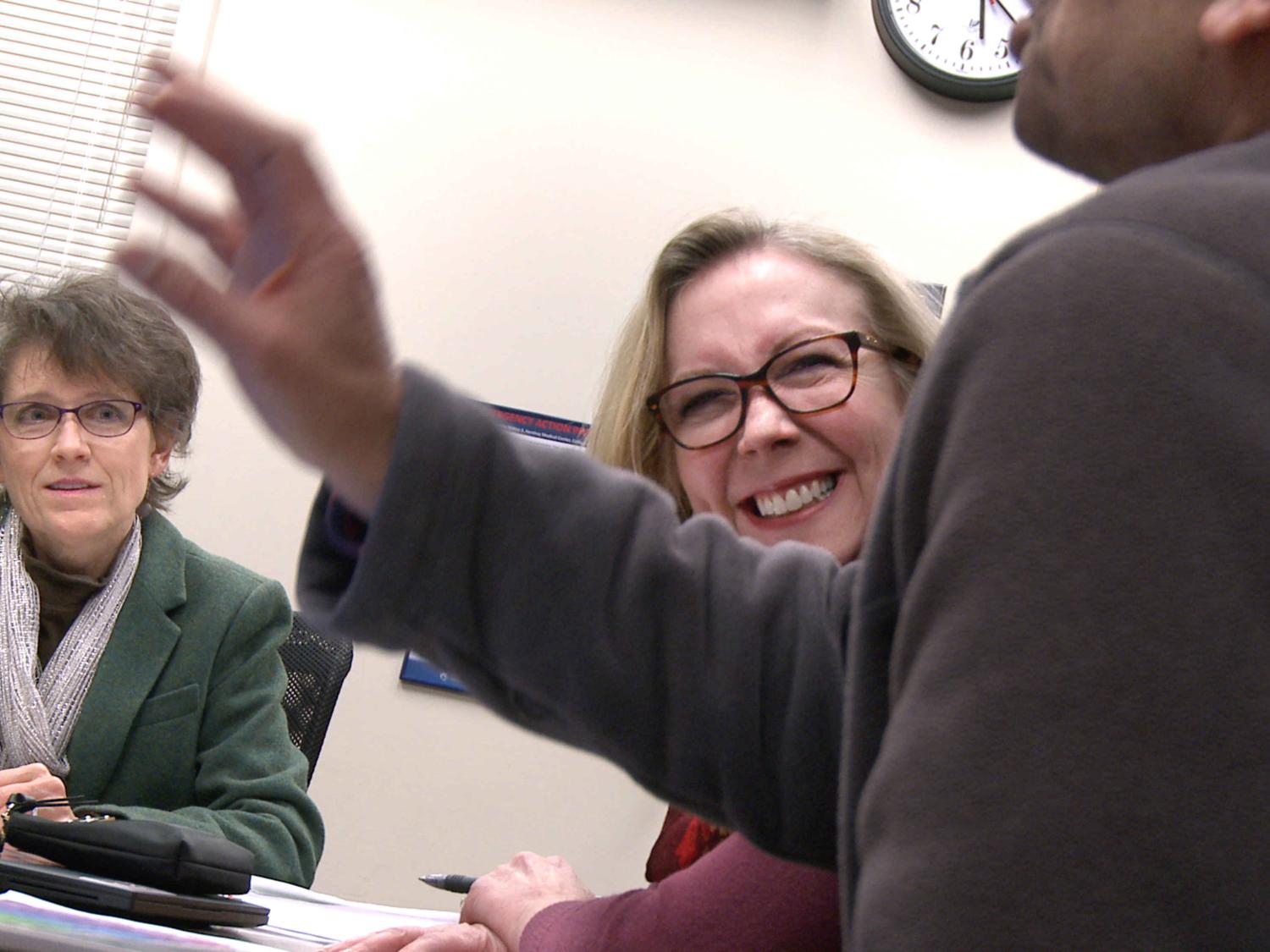 Physician Writers Group founder Kimberly Myers and Dr. Eileen Moser look at Dr. Jose Stoute as he presents an original work during a workshop in February. They are seated at a table with papers and laptops on it.