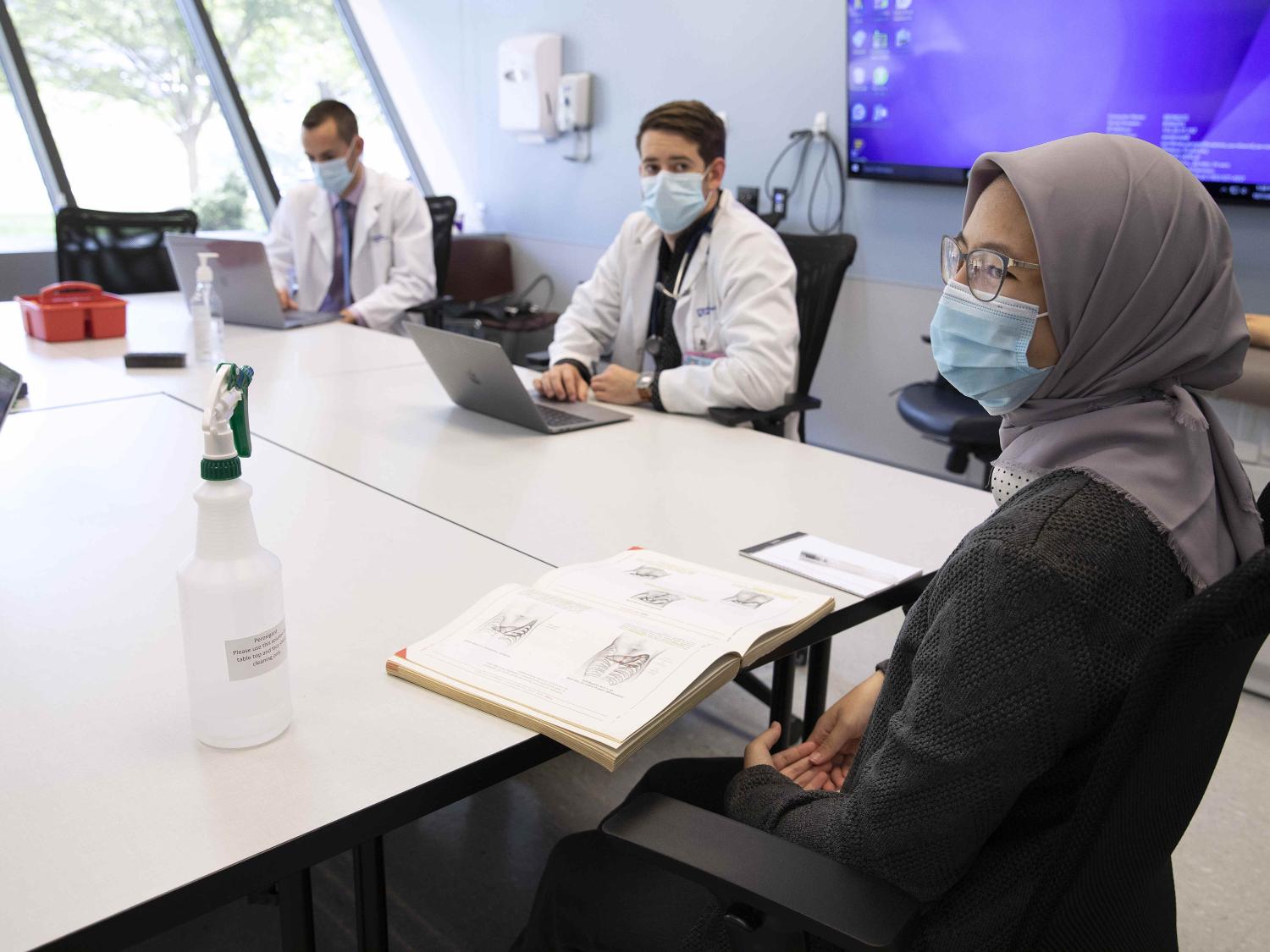 A female medical student wearing a surgical mask and head covering is seated at a conference table with an open textbook in front of her. In the background, two male students, also wearing surgical masks, are seated at adjoining conference tables.