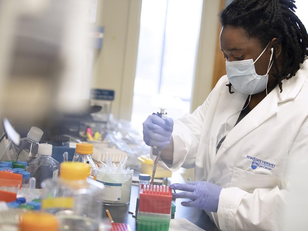 Jasmine Geathers is working in a lab at Penn State College of Medicine. She is wearing a white coat with the Penn State Hershey logo on it, a face mask and gloves. She is holding an instrument in a container of test tubes.