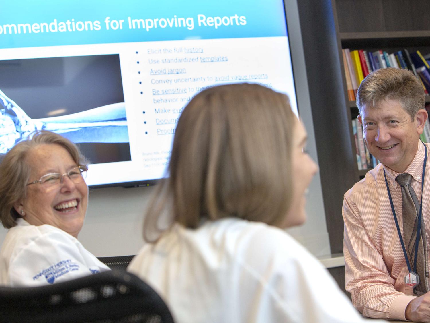A man leans on a table as he talks with two women, who are seated and either smiling or laughing. A book shelf and projector screen are in the near background.