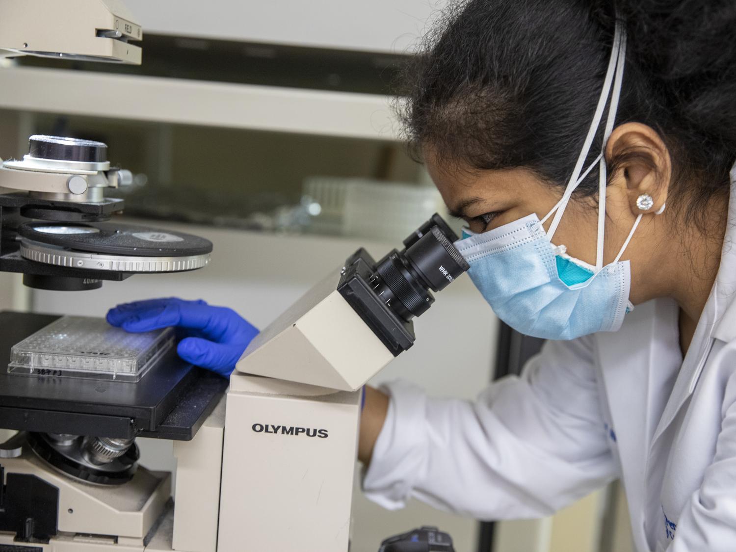 A woman wearing a mask and lab coat looks into a microscope.