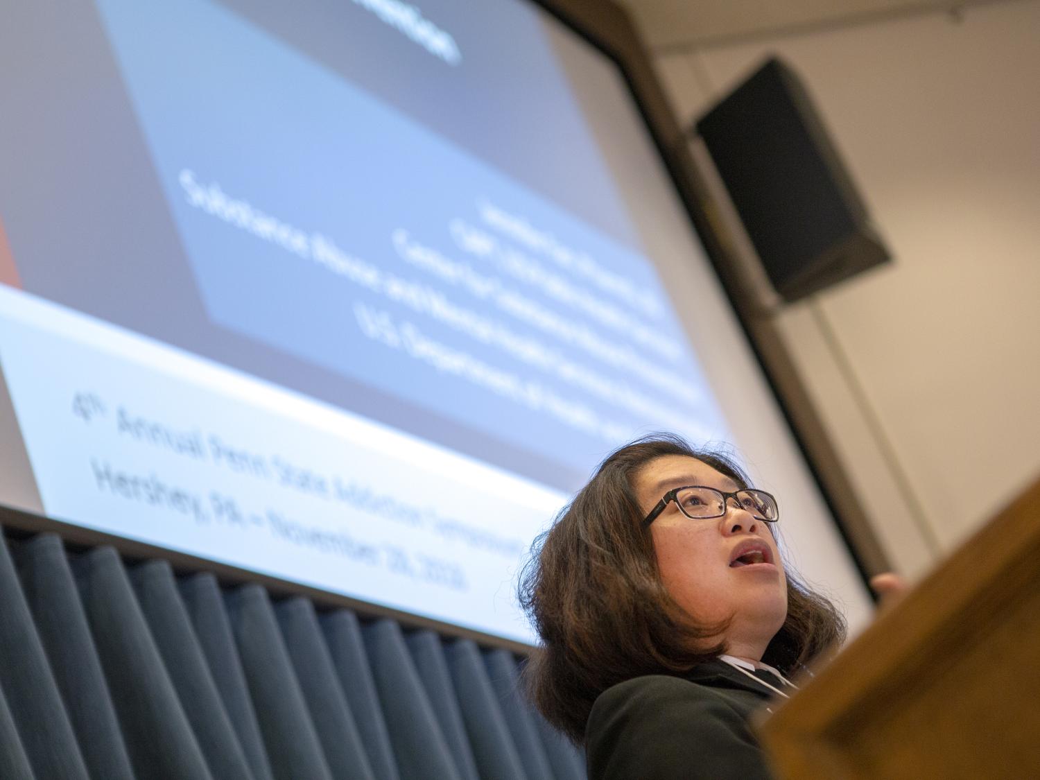 A woman stands at a lectern delivering a speech. A screen with a PowerPoint presentation is in the background.