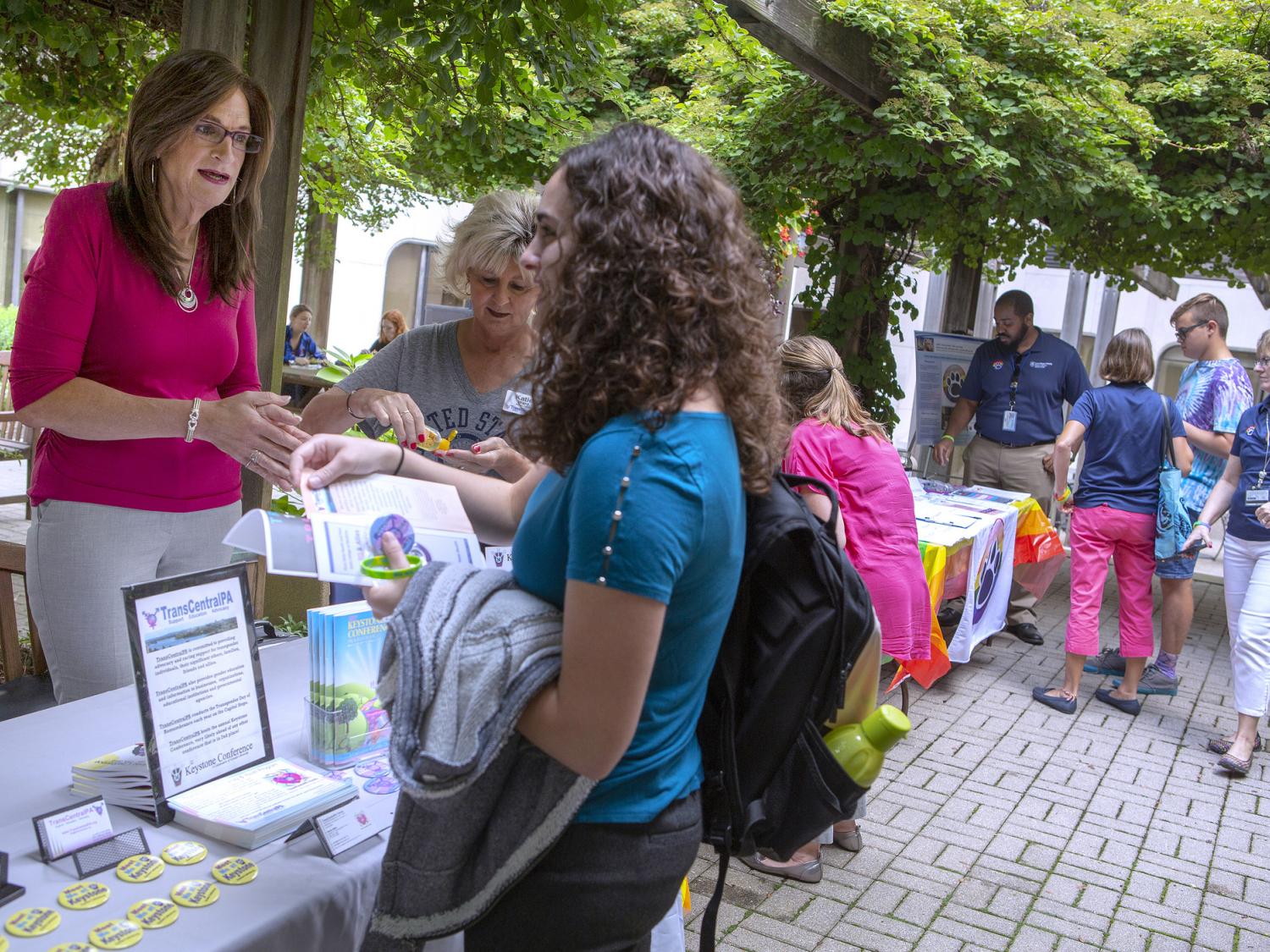 Two women stand across a table from each other, talking. Several signs and brochures are on the table. They are outside. Several people are in the background.