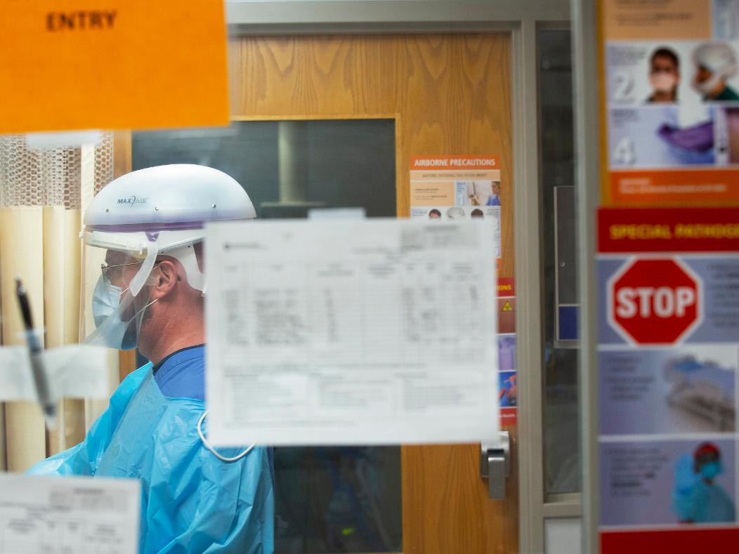A man dressed in personal protective equipment including a gown, mask and face shield prepares to enter a patient room. Signs indicating the use of personal protective equipment are posted around him. 