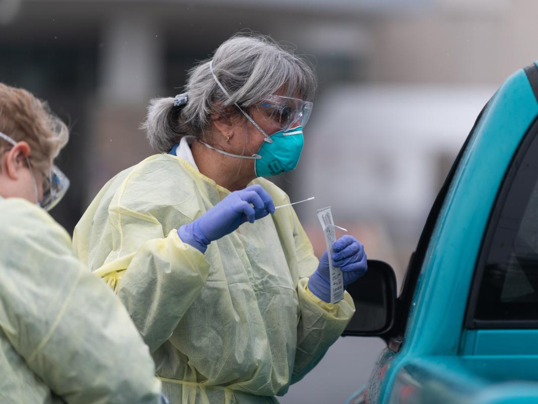 Two women in personal protective equipment, including gowns, masks, goggles, and gloves, stand alongside a vehicle, speaking to the driver, who’s not visible. One worker holds a swab.