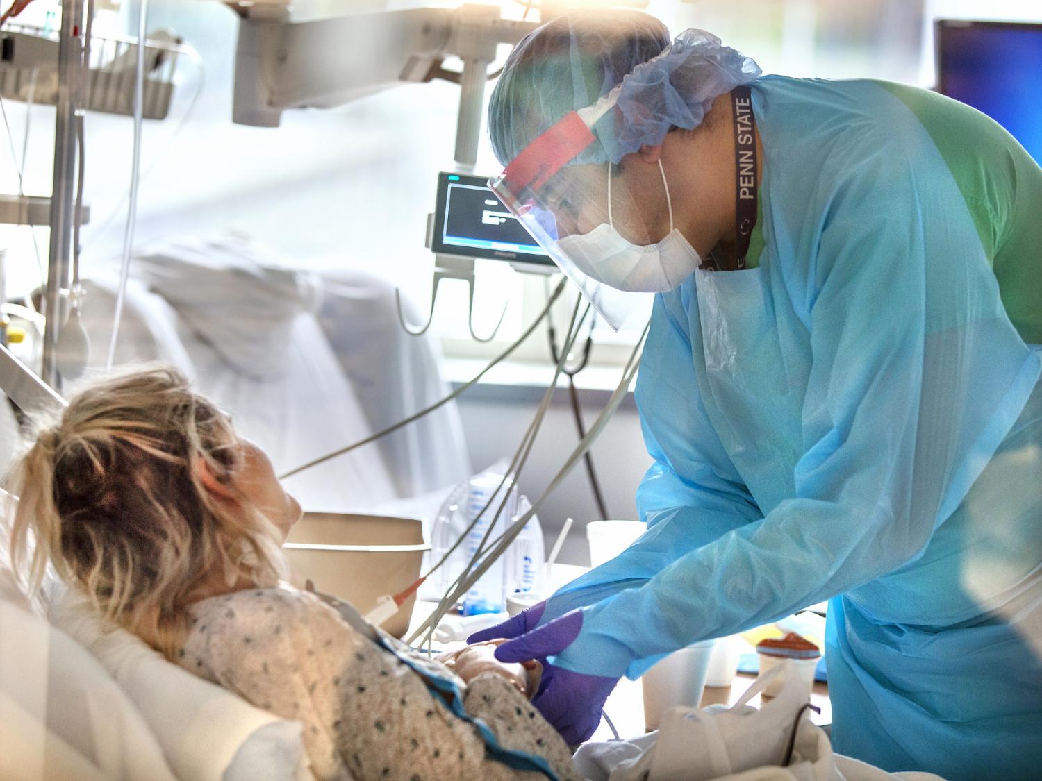 A caregiver wearing a gown, mask, face shield and gloves, holds the hand of a female patient who is in a hospital bed. A monitor and window are among items in the background.