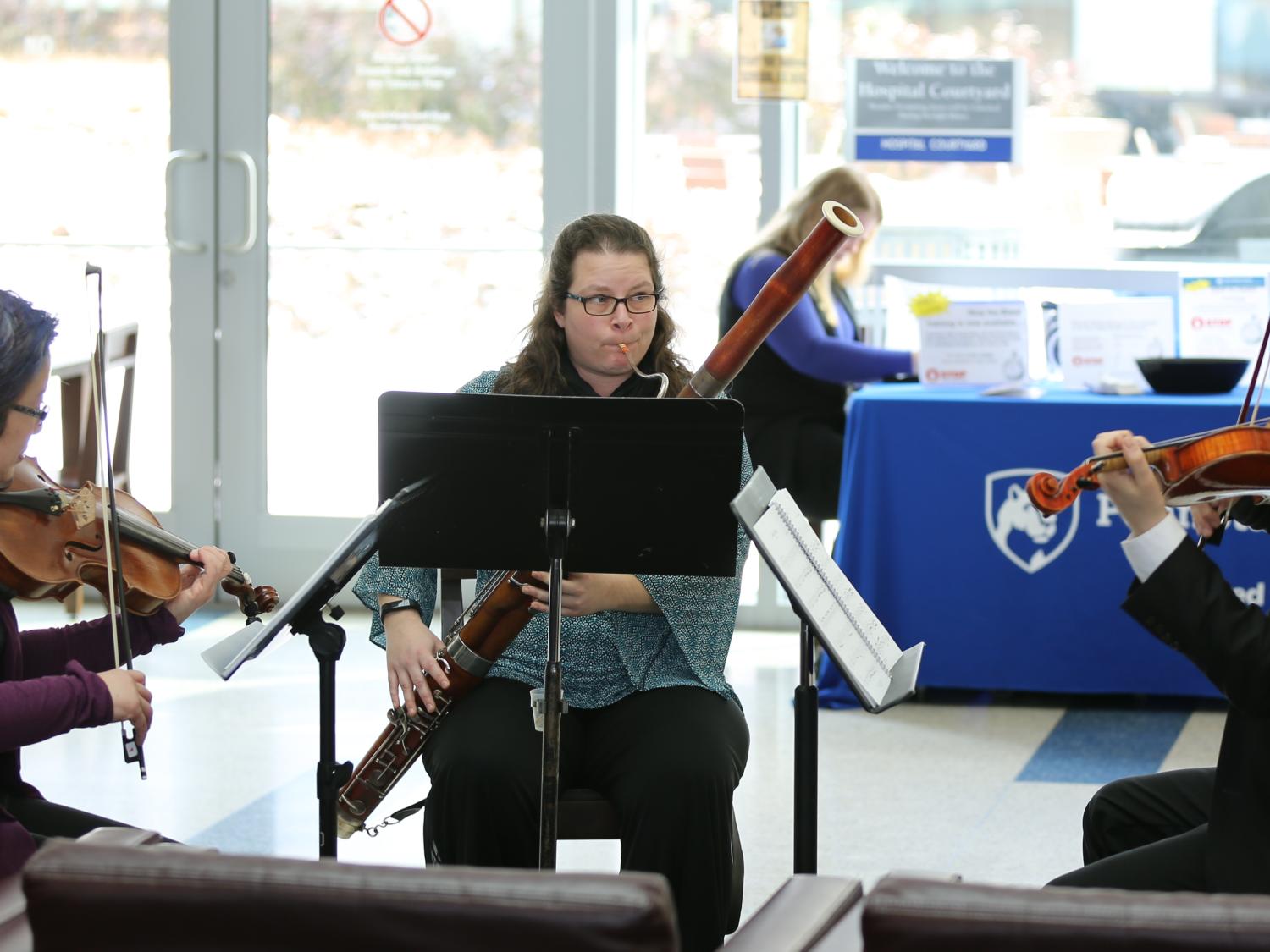 A trio of musicians - a violinist at left, bassoonist at center and violinist at right - play their instruments. In the background are windows and doors that open to a courtyard.