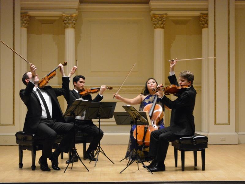 Four musicians sit at music stands and hold their bows above their string instruments.