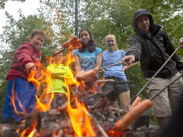 A view through the middle of a campfire as four children and one adult stand around it. Trees are in the background.