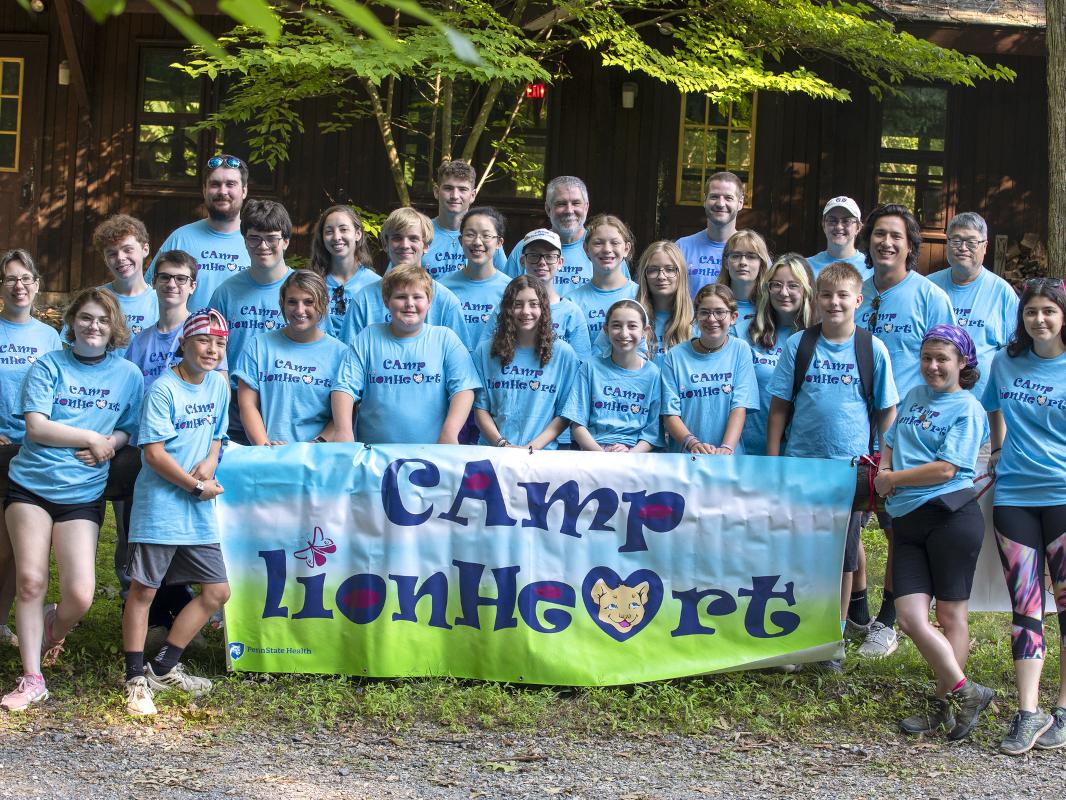 A group of teens and a few adults pose behind a large sign that reads “Camp Lionheart.” A building and some trees are in the background.
