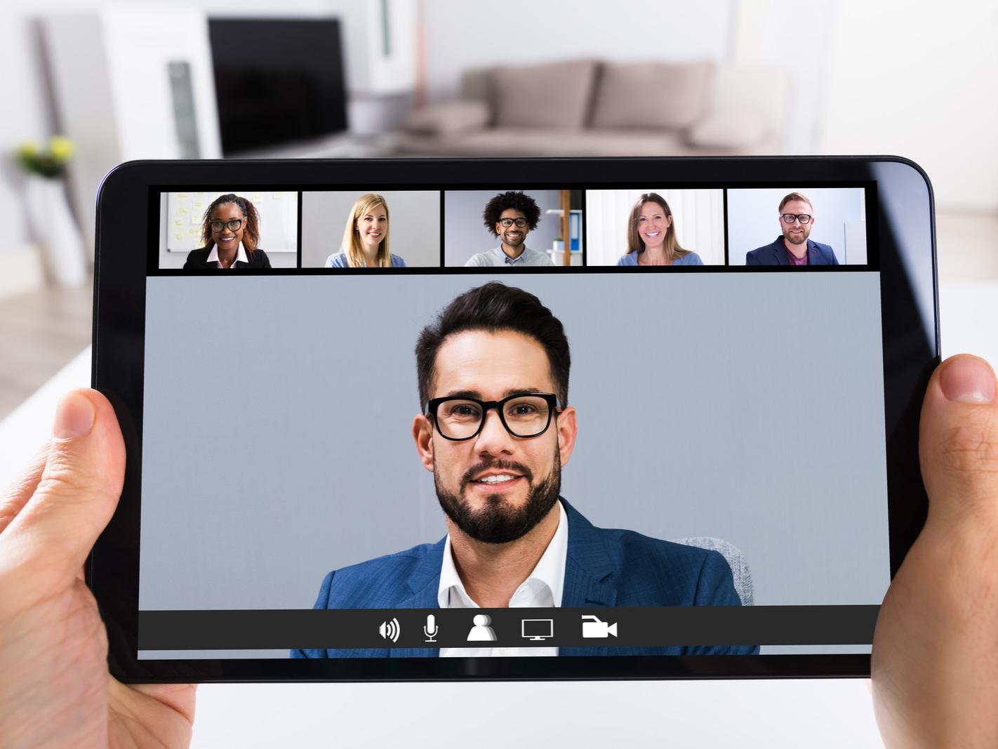 A person holds a tablet computer as it displays faces of Zoom-style meeting participants