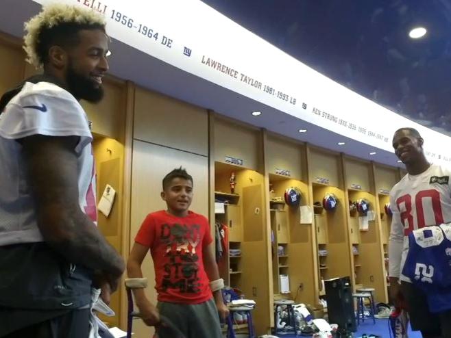 Deven Jackson stands in the middle, talking with two NFL football players (Odell Beckham and Victor Cruz), who are on either side of him in the Giants' locker room.