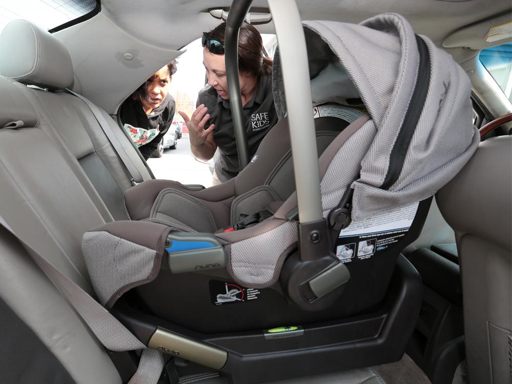 The view from the back seat of a car, across a rear-facing infant car seat, as two women peer in through the opposite side door, talking.
