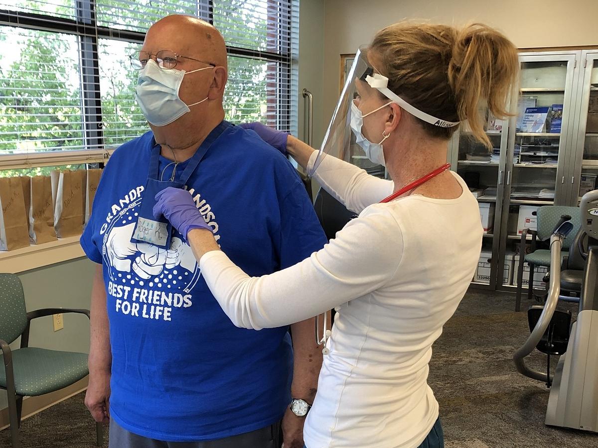 An exercise physiologist touches the back of a male patient as he stands in a cardiac rehabilitation room. The man is wearing a heart monitor and face mask. The exercise physiologist is wearing a face mask and face shield.