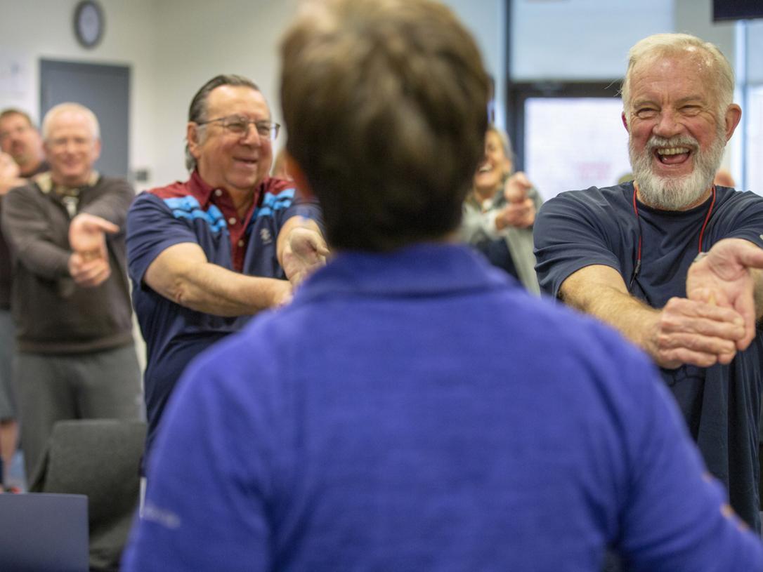 Hank Shipman smiles broadly as he stretches his arms out in front of himself and bends down his fingers. He wears a T-shirt and has a beard and moustache. Other rehab patients are in the background. The instructor is seen from behind.