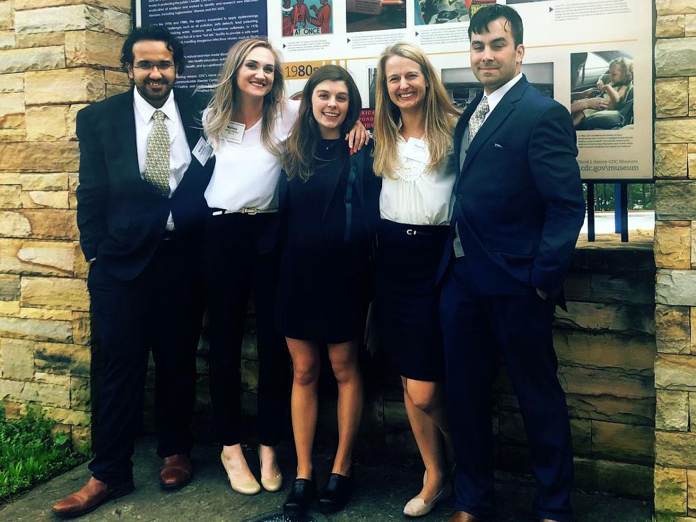 Five people – two men and three women – wearing semi-formal/business attire stand outside in front of a sign with a stone base.