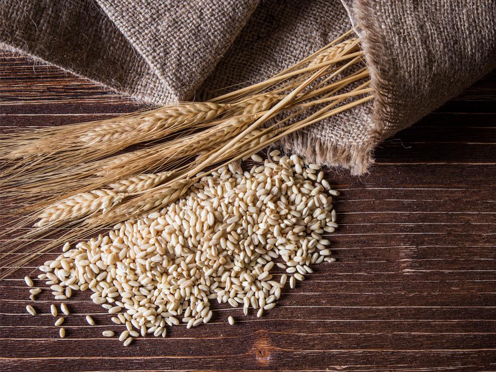 A burlap sack rests on a wooden surface. Wheat and other grains are spilling out of the top of the sack.