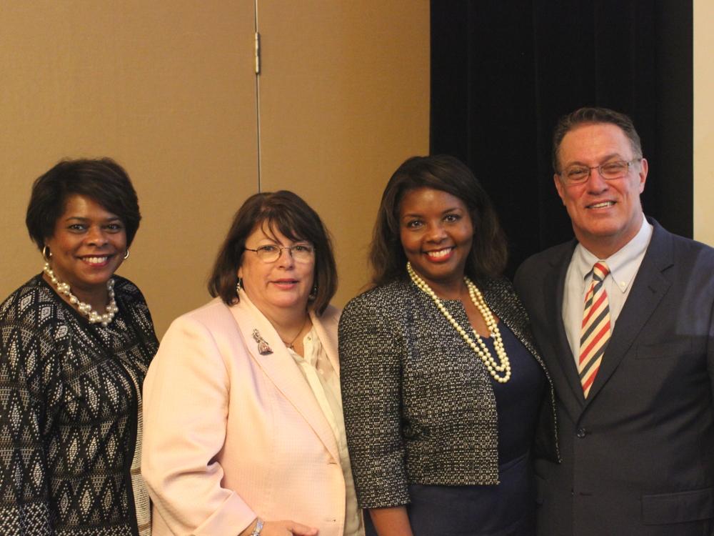 Four people – three women and one man – pose for a photo, in front of a beige and black backdrop.