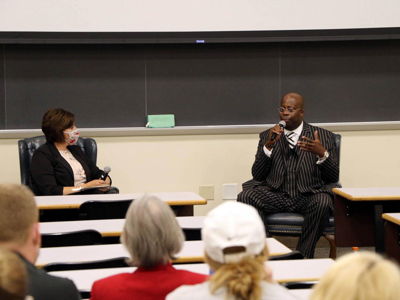 Man with microphone answering a question in a classroom with female moderator seated next to him.