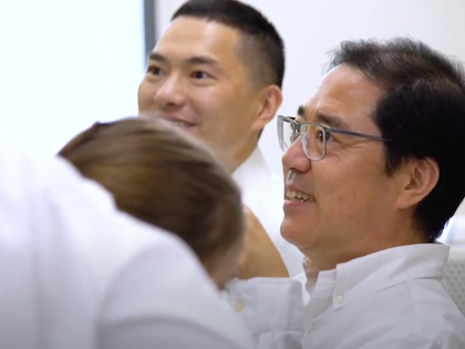 Man in white lab coat smiling while looking at data with another man in background and woman in foreground.