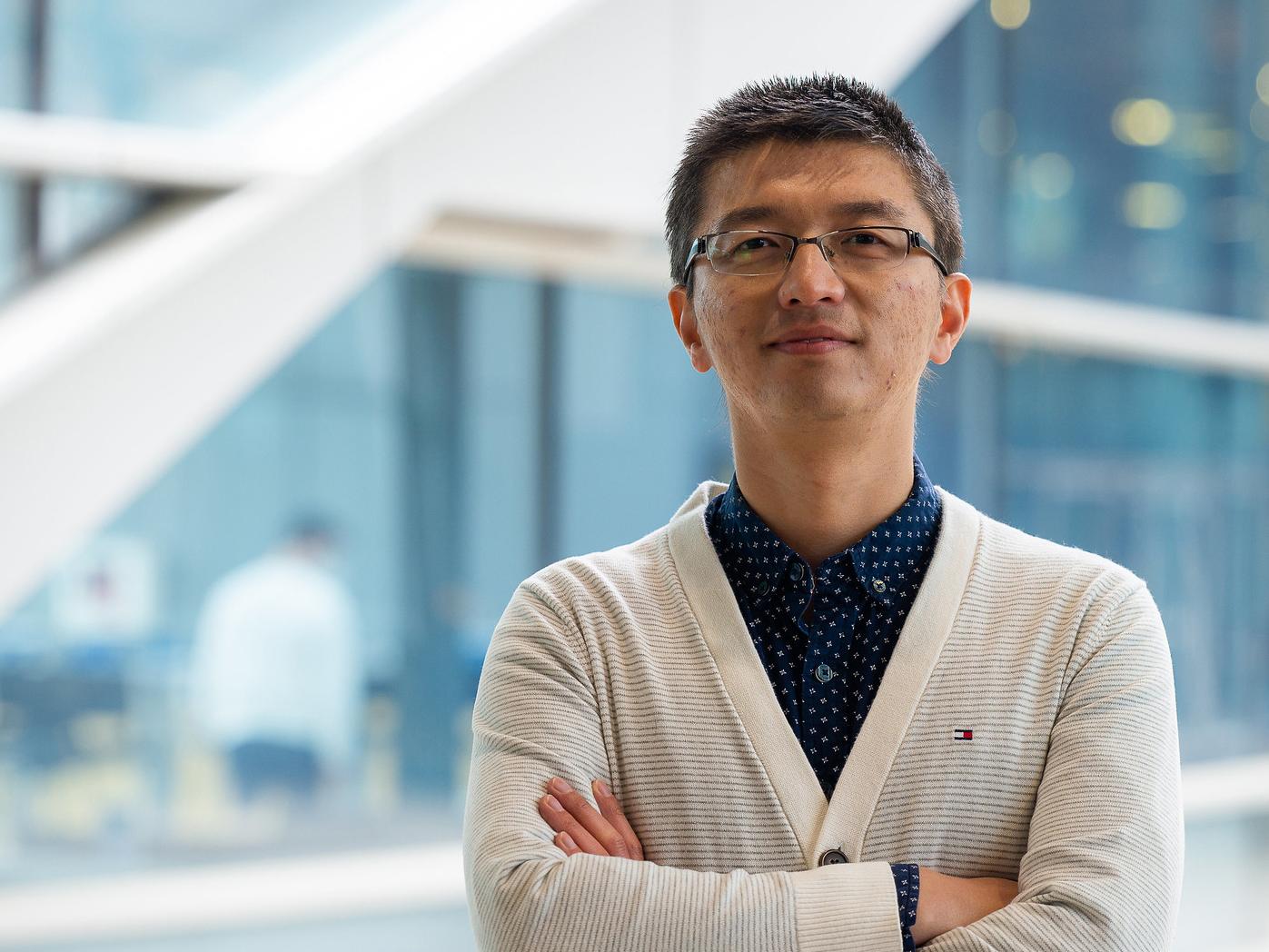 A man wearing glasses and a white cardigan poses for a photo in front of a window.