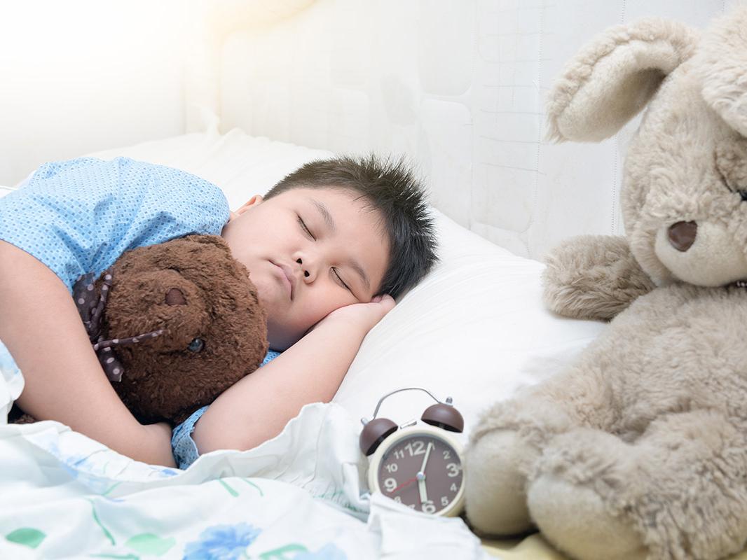 A child sleeps in a bed holding a teddy bear, while laying next to a larger teddy bear.