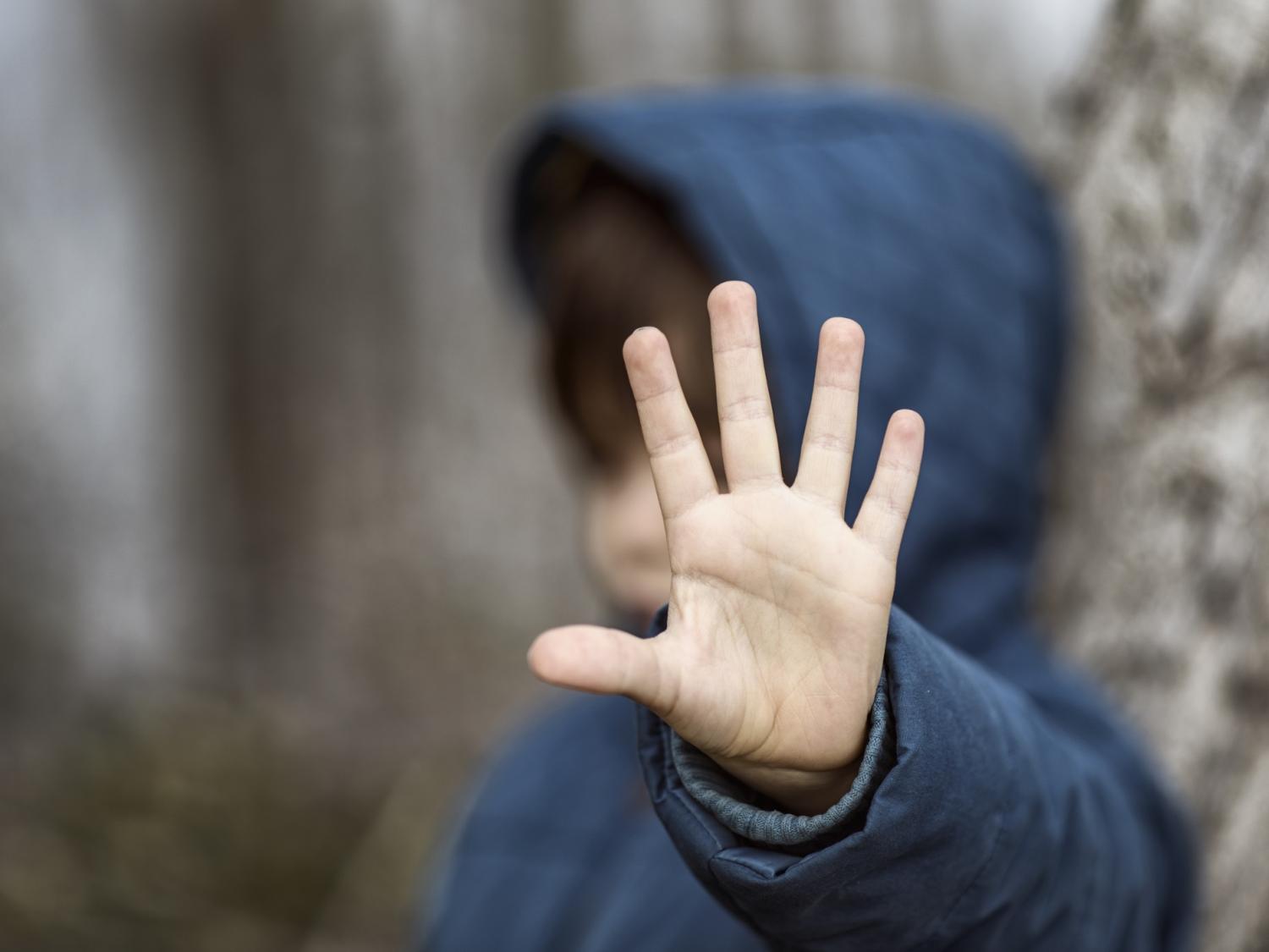 A child wearing a sweatshirt with a hood is seen out of focus and puts his hand up toward the camera.