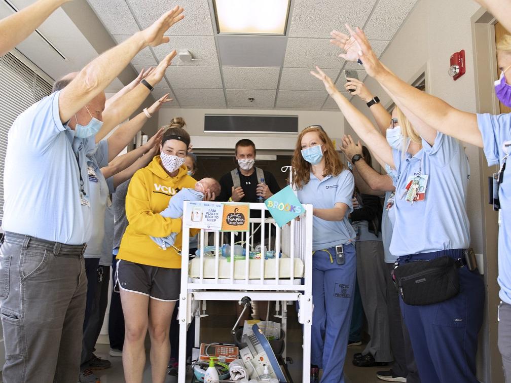 Men and women in polo shirts and surgical masks hold their hands over their heads, forming a tunnel for a man, a woman, a baby and another health care worker rolling a crib down a hallway.
