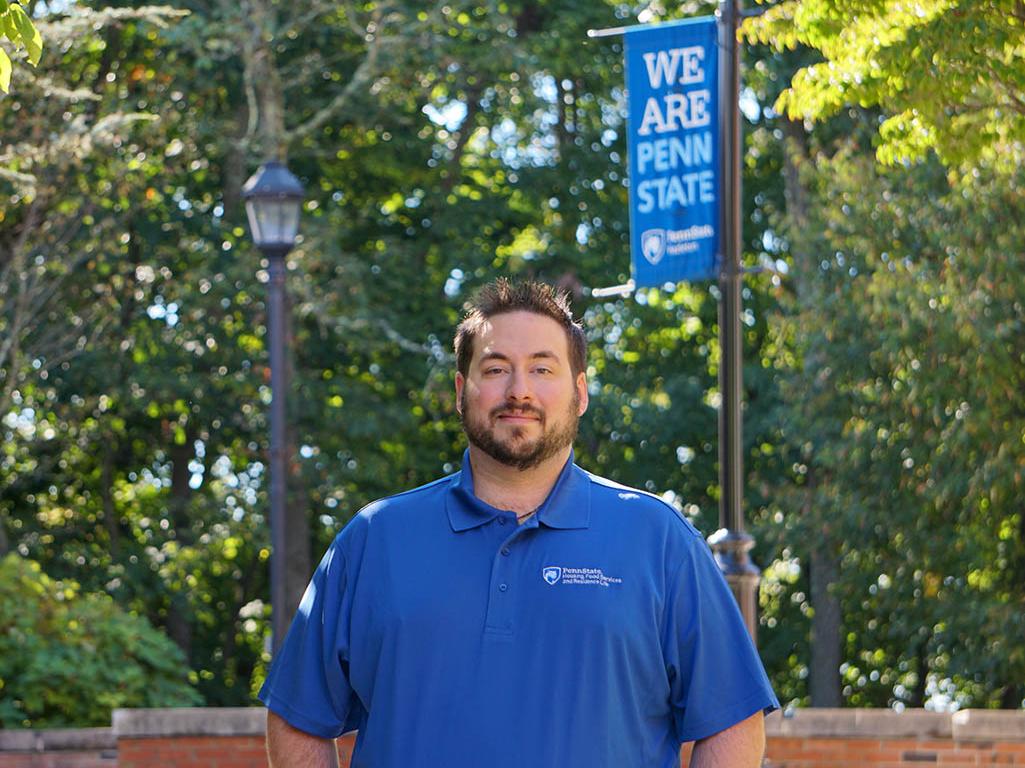Man in blue polo shirt standing in front of trees and Penn State Hazleton banner hanging from light post.