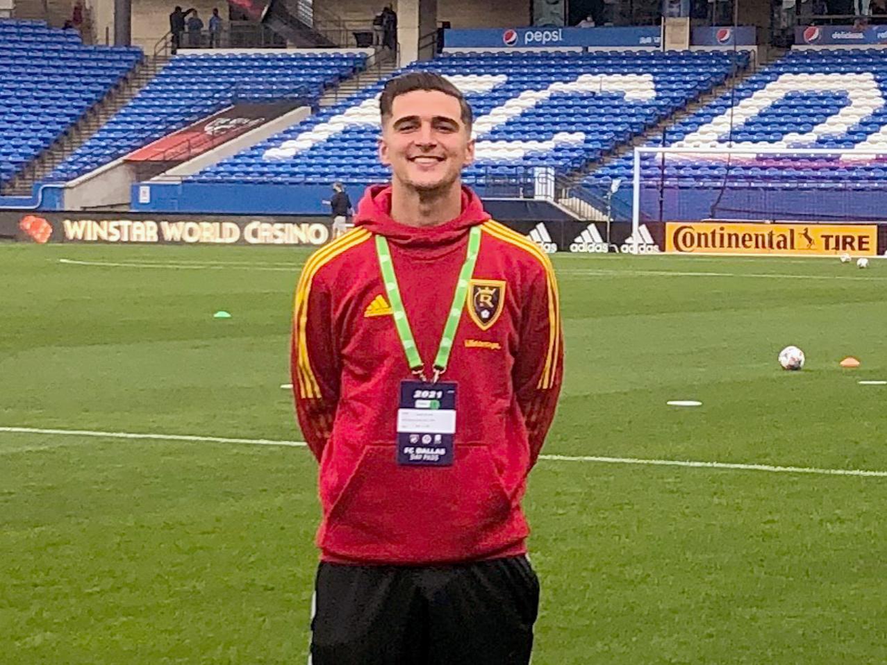 Christian Peters stands on a green soccer field wearing a red sweatshirt and black shorts.