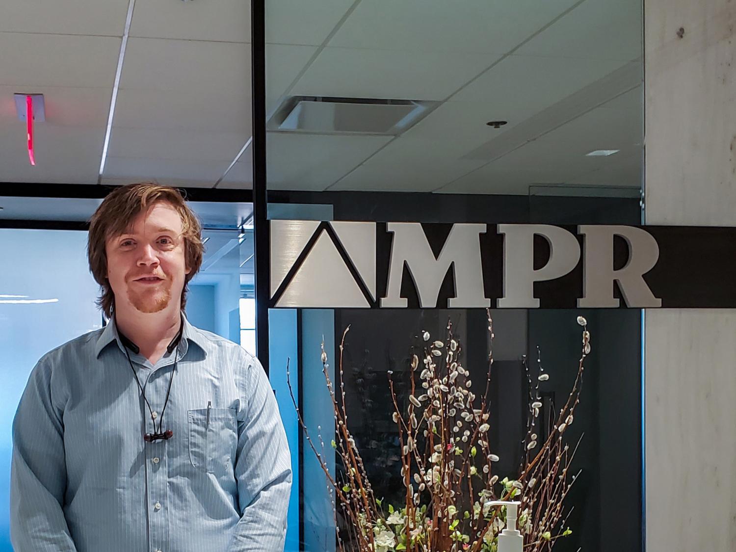 Christopher Hile standing in front of a glass wall in an office with an MPR sign behind him. 