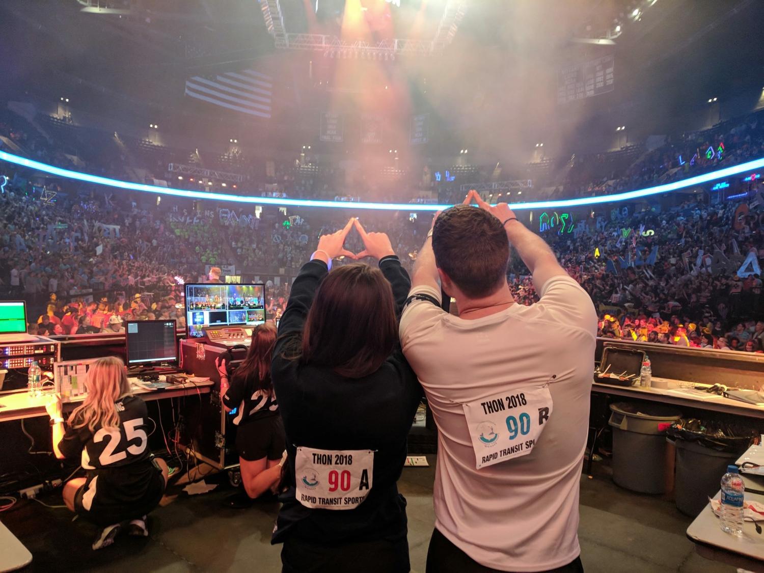 A view from behind as two people stand side-by-side on a stage facing thousands of people in an arena. Both people have their arms in the air, and their fingers in a ‘diamond’ formation.