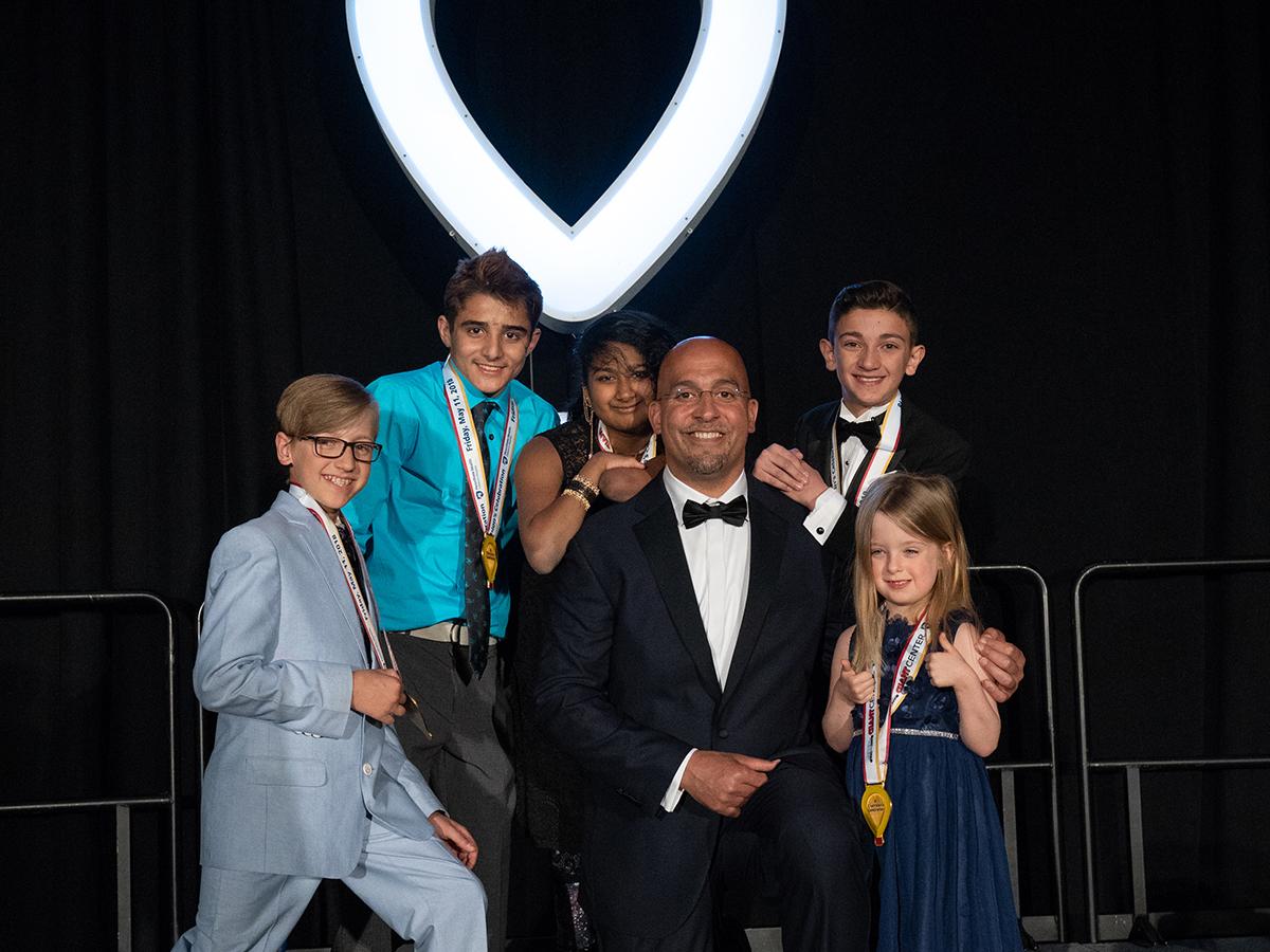 Penn State Football Coach James Franklin, wearing a tuxedo, kneels on one knee. Around him are five young children. An illuminated CMN logo is in the background.