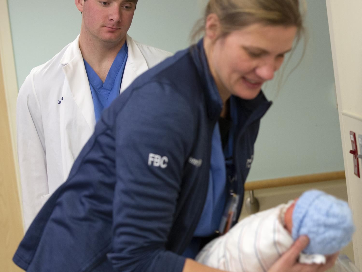 Colin Hayes watches as Jenny Meincke, a registered nurse at Penn State Health St. Joseph Medical Center, leans over while holding a newborn baby. Hayes is wearing a white coat and scrubs. Meincke is wearing a jacket and scrubs.