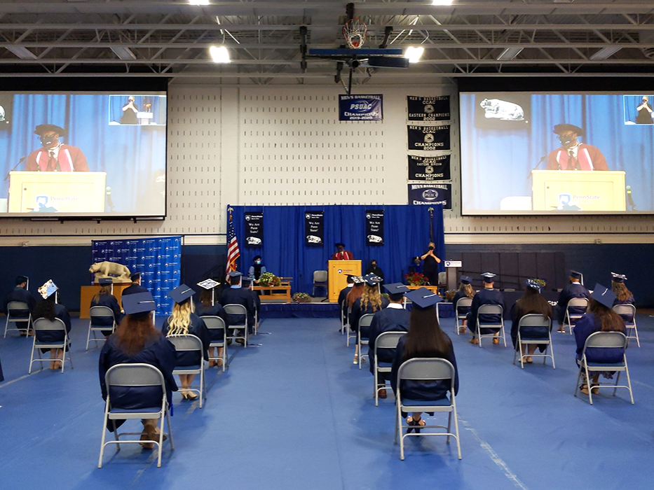 Students in regalia in the campus gym listening to remarks from the chancellor