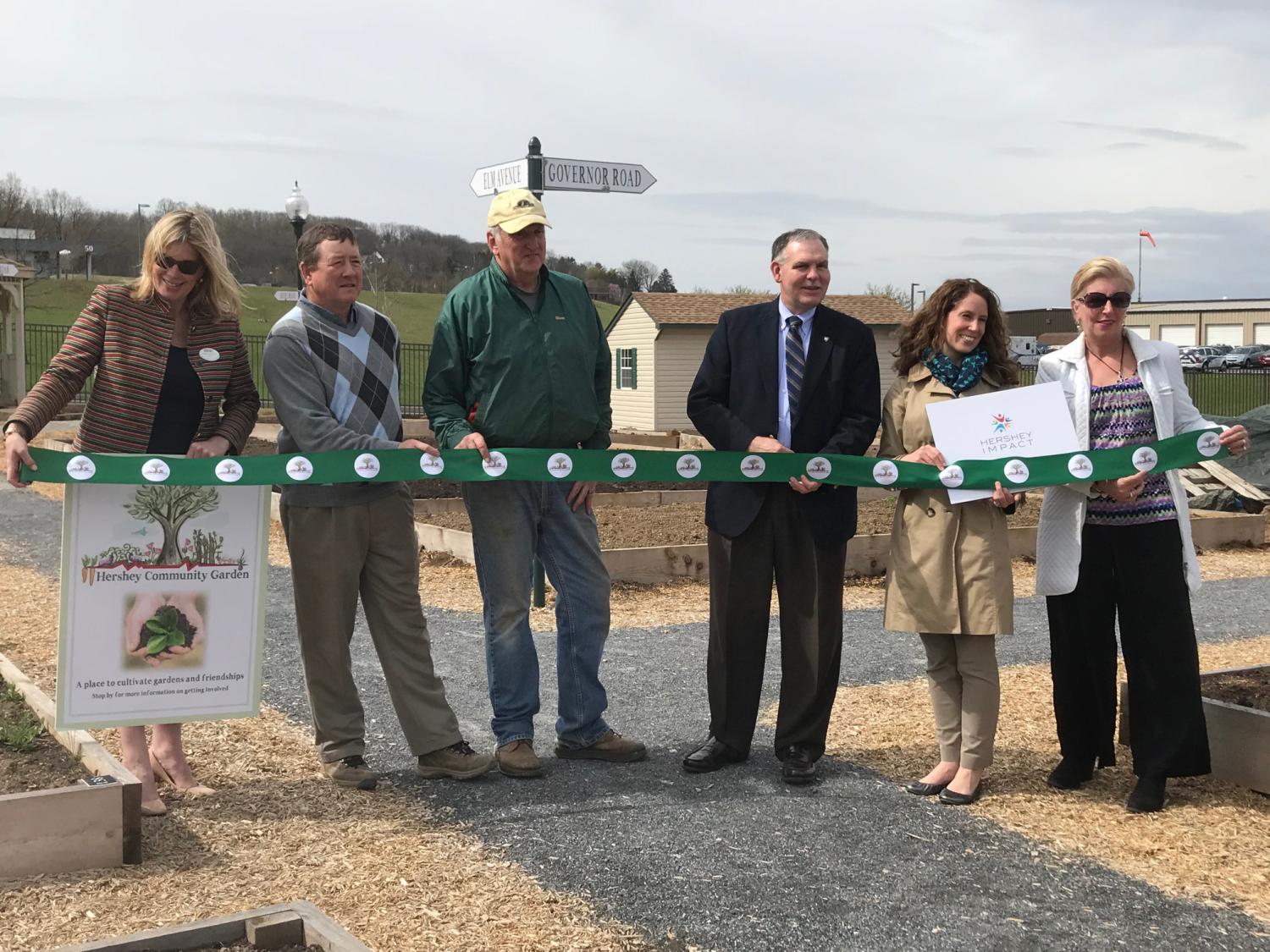 Six people – three women and three men – stand behind a green ribbon which they hold while posing for a photo. They are outside, with garden plots and grass in the background.