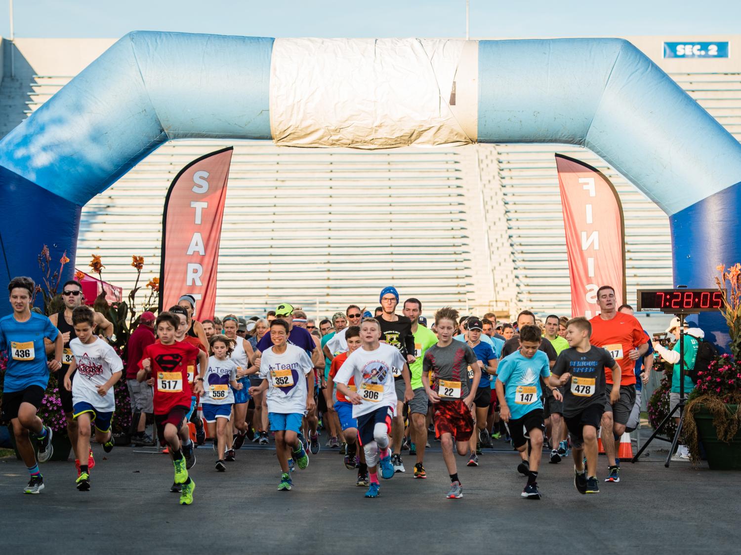 A group of children and adults in running clothes run through a large blue inflatable arch, underneath which there are signs reading 'start' and 'finish.'
