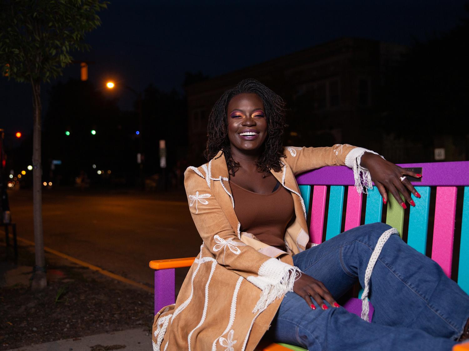 Woman in tan jacket sitting on multicolored park bench at night.