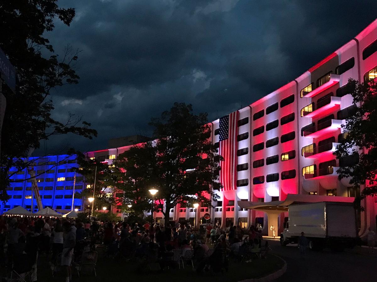 At night, the Hershey Medical Center's signature Crescent building is lit up in red, white and blue lights. Several people are sitting in the foreground.